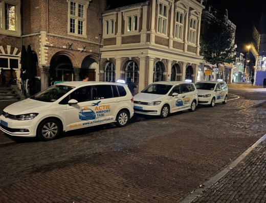 Drie witte taxi's van Actie Airport Taxi met bedrijfsbelettering achter elkaar geparkeerd in een stadsstraat.